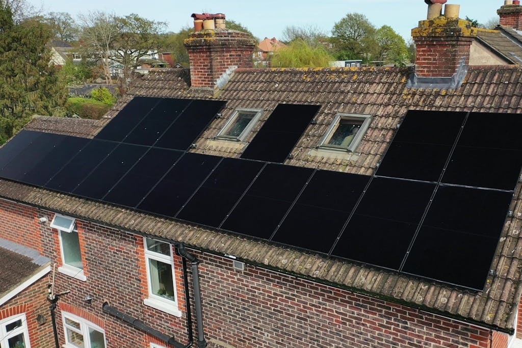 Black solar panels on a brown tiled roof, under brick chimneys and a blue sky