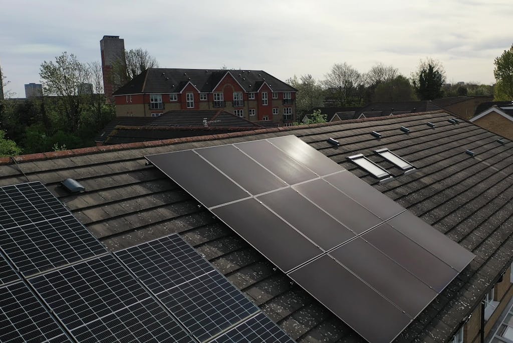 Black solar panels next to blue solar panels on a dark roof near another large house, under a light blue sky