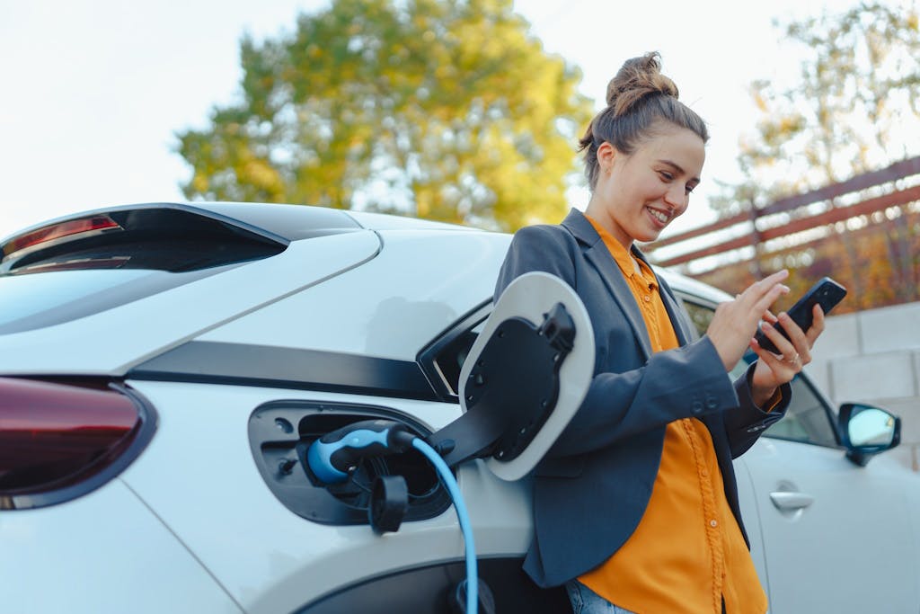 Person with a smartphone leaning against an electric car with a charger in it, looking at their smartphone