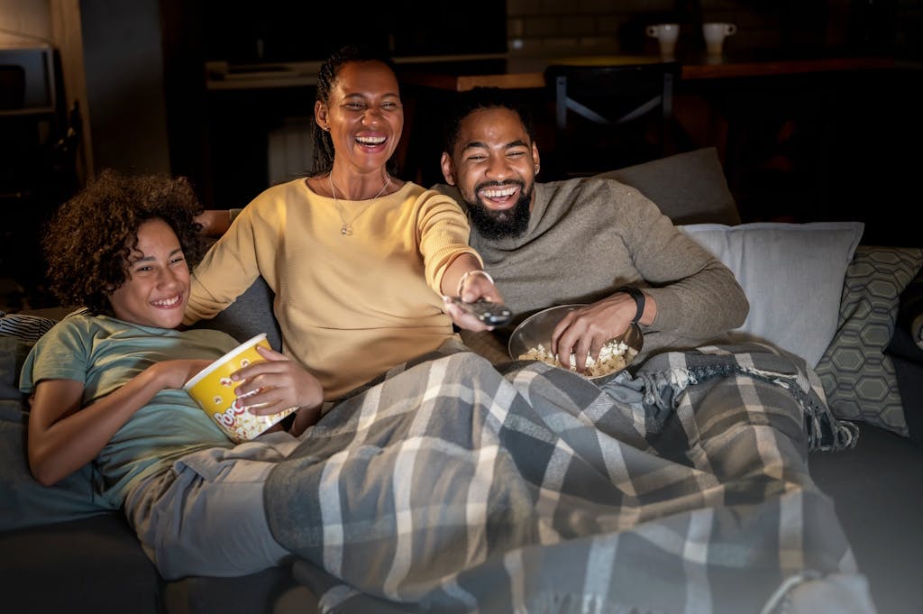 A woman, man, and adolescent laughing on the sofa while bathed in TV light. They're covered in a blanket, eating popcorn