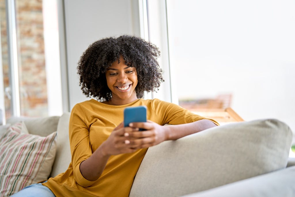 Woman of colour holding a smartphone on a cream sofa, with a bright white wall in the background