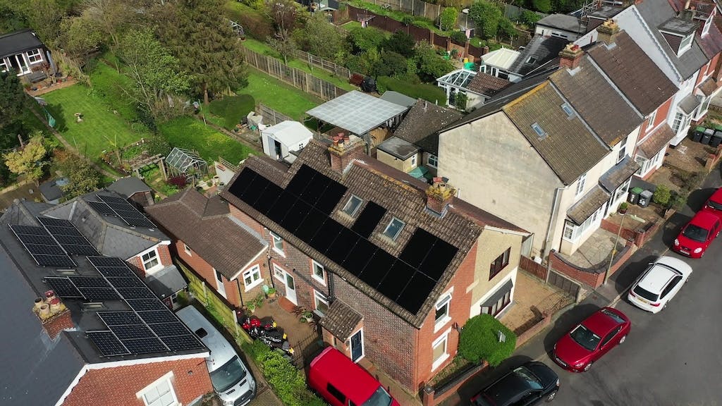 Aerial view of an array of black solar panels on a rooftop, cars parked along the road out front, green garden at the back