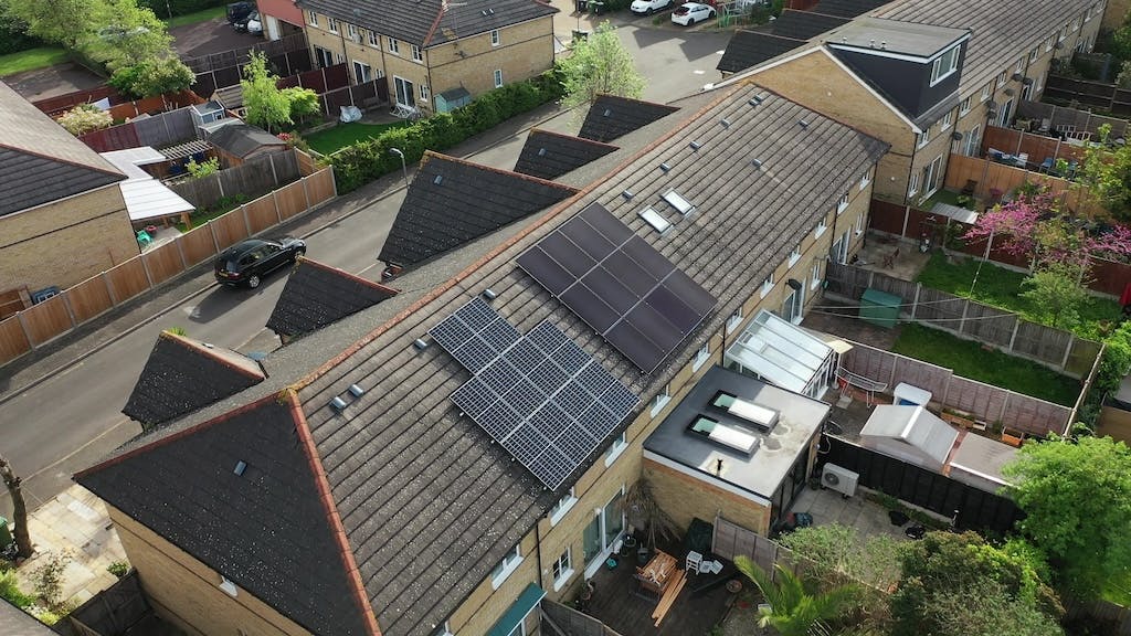 Aerial shot of a 12-panel solar panel system on a rooftop of a terraced house, lots of fenced gardens on one side and road on another