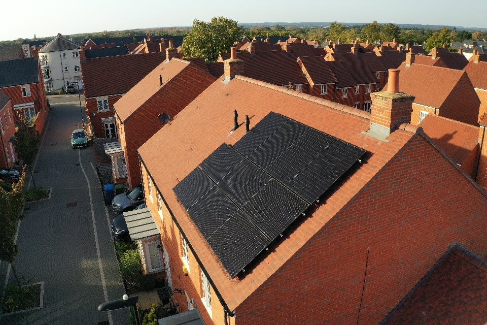 A bird's-eye view of black solar panels on a terracotta roof of a detached house, near other houses and trees, under a pale blue sky