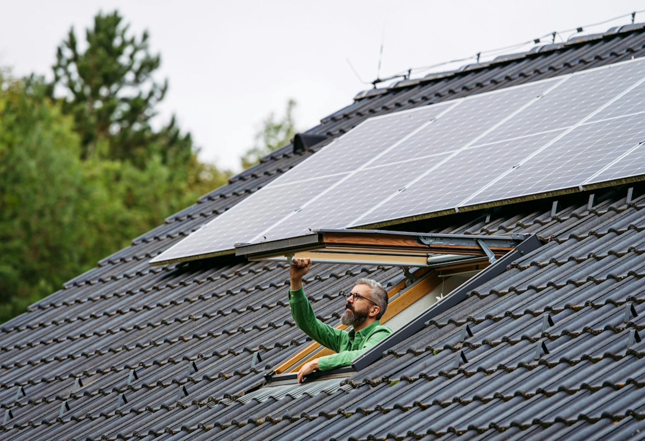 a dark grey roof with black solar panels and a man in green opening a skylight