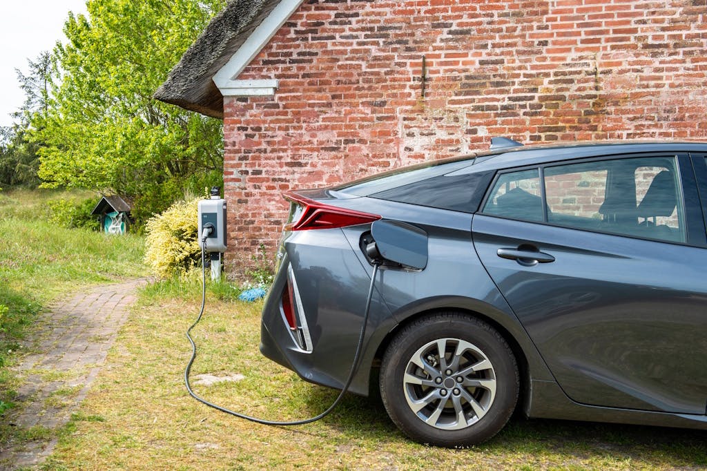 A charger connected to a dark electric vehicle in front of a brick wall, by a garden