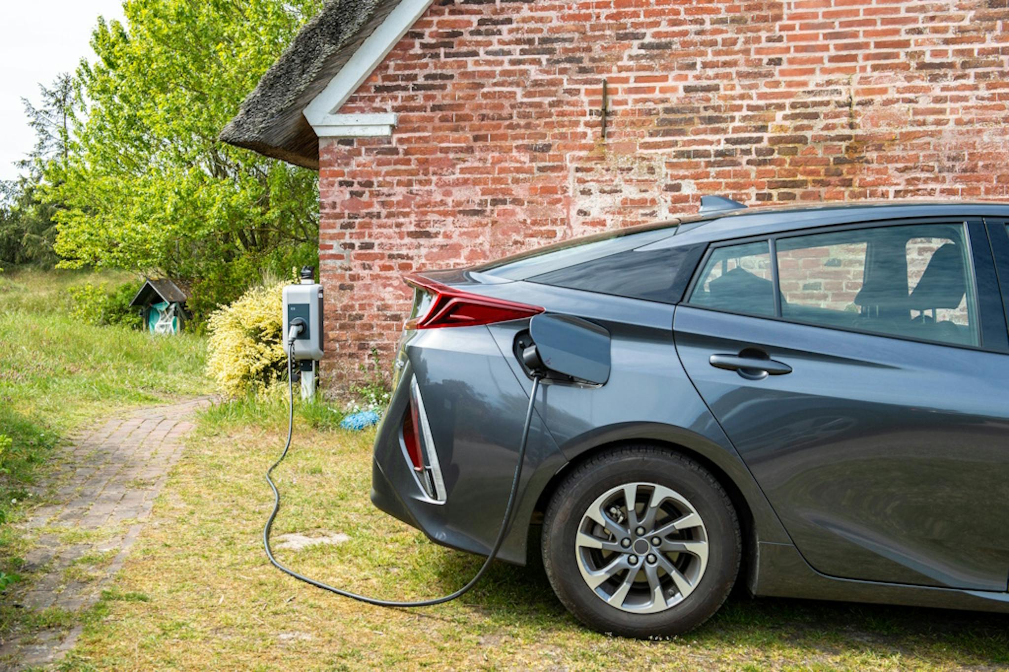 A charger connected to a dark electric vehicle in front of a brick wall, by a garden