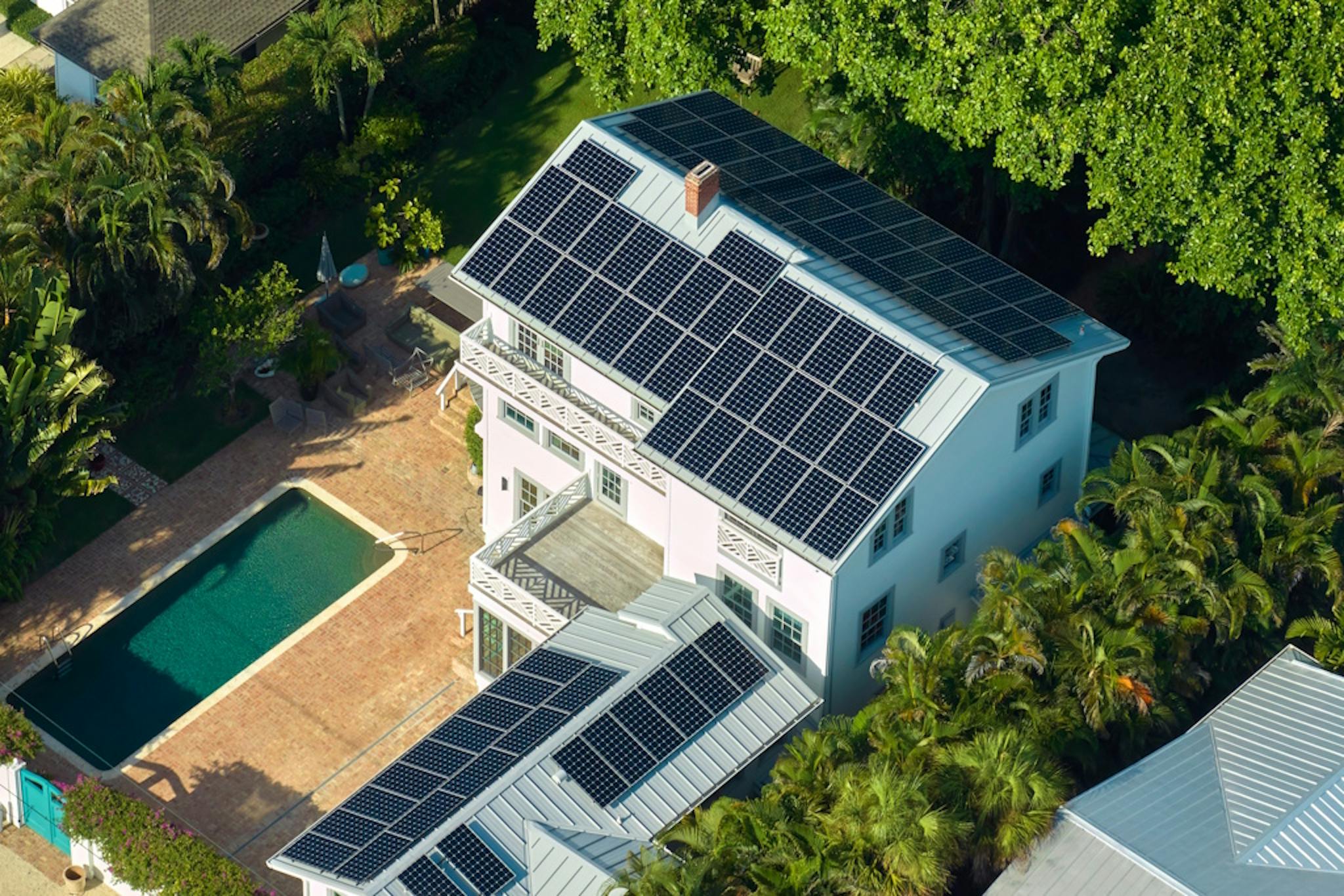Aerial view of black solar panels on the roof of a large home and shed, next to a swimming pool