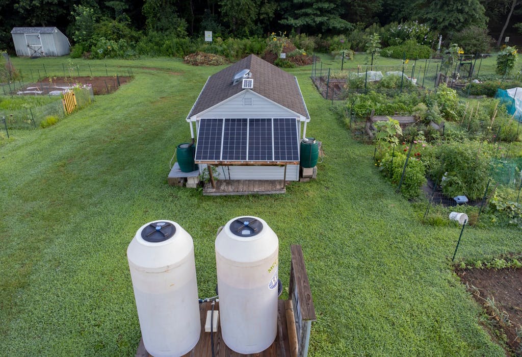 a small home with two white storage cylinders and solar panels, in the middle of a garden, next to an allotment