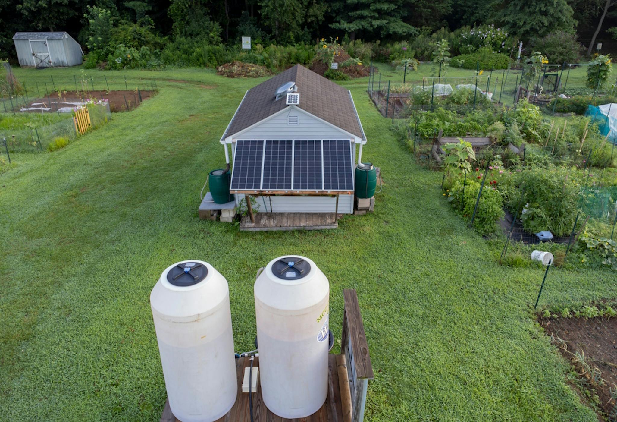 a small home with two white storage cylinders and solar panels, in the middle of a garden, next to an allotment