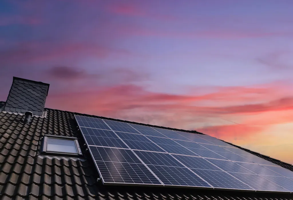 black solar panels on a roof by a Velux window, under a sunset sky