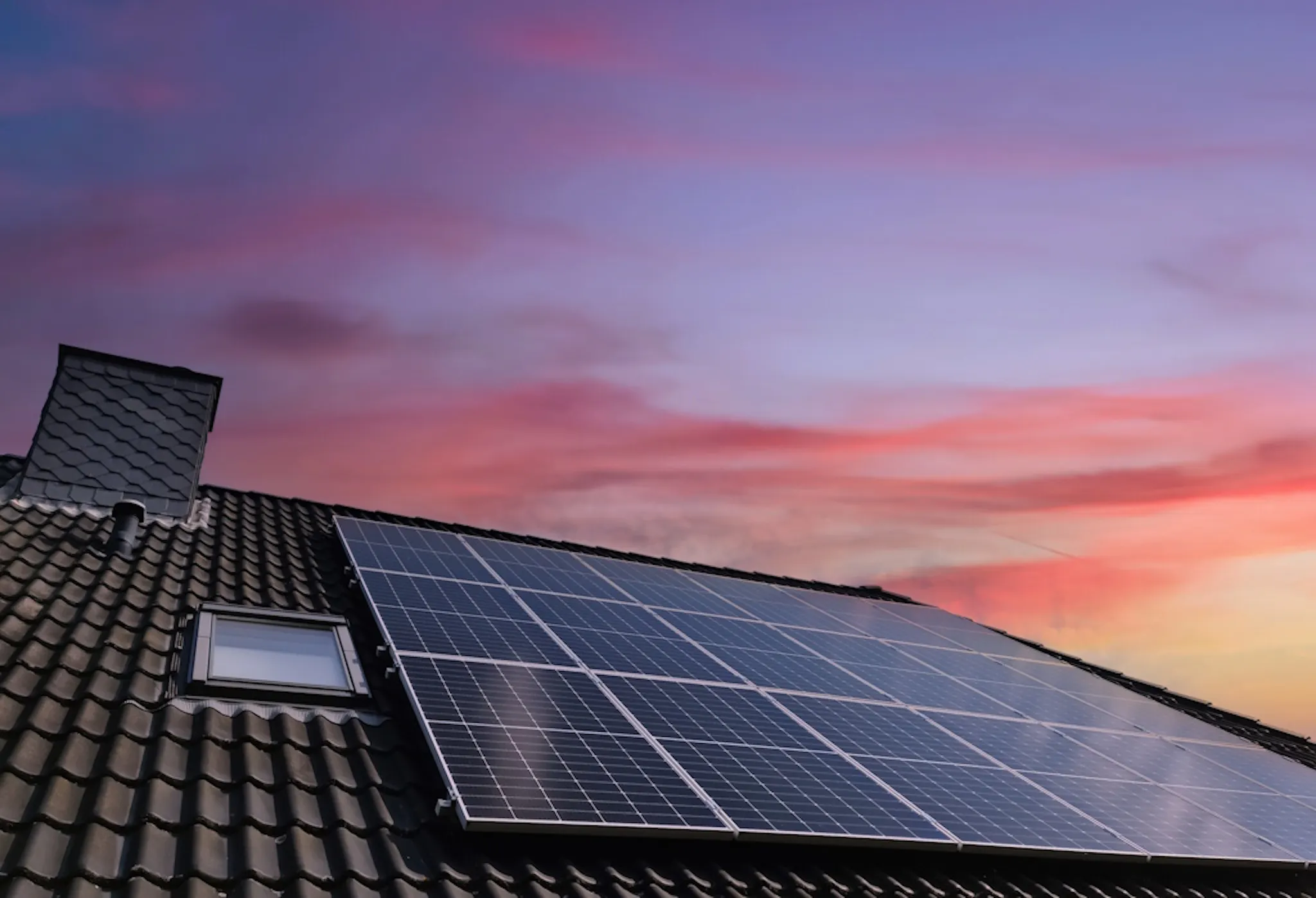 black solar panels on a roof by a Velux window, under a sunset sky