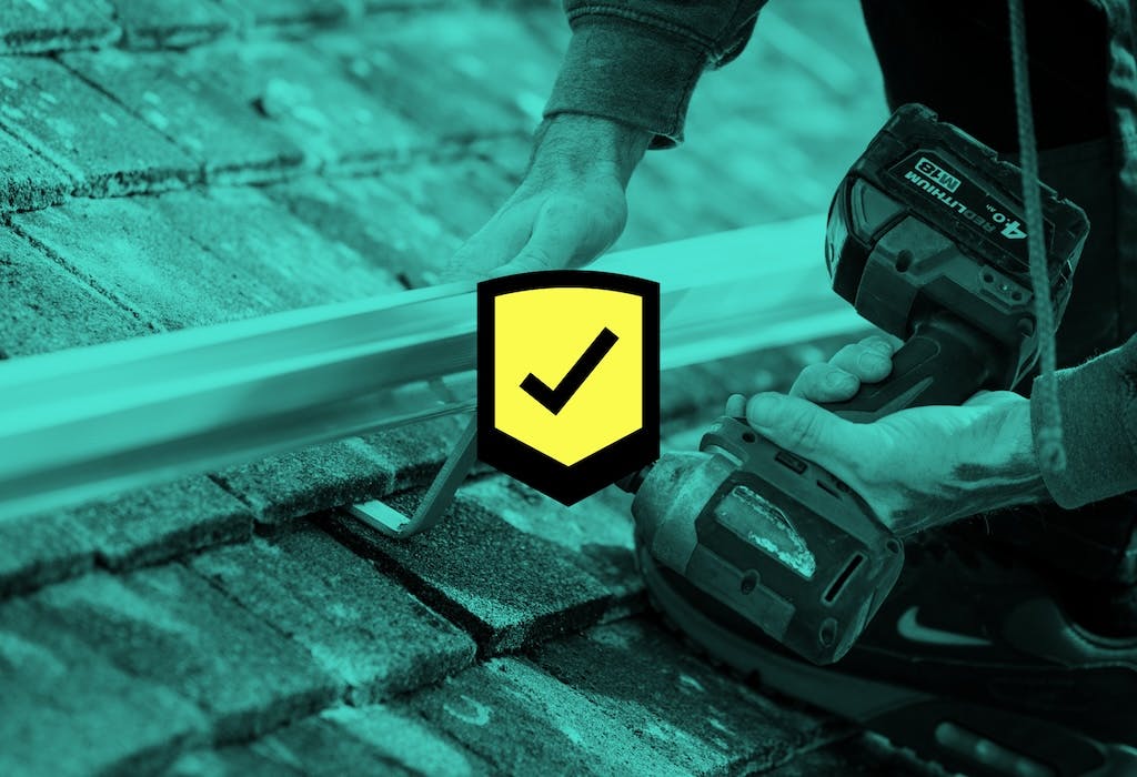 Solar panel installer drilling into a mounting bracket on a rooftop, blue filter overlaid, black tick in a yellow box in the centre foreground