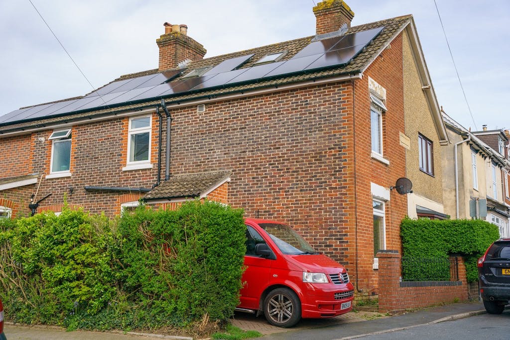 Black solar panel array on top of a house in the UK, red van parked in the driveway beside a green hedge