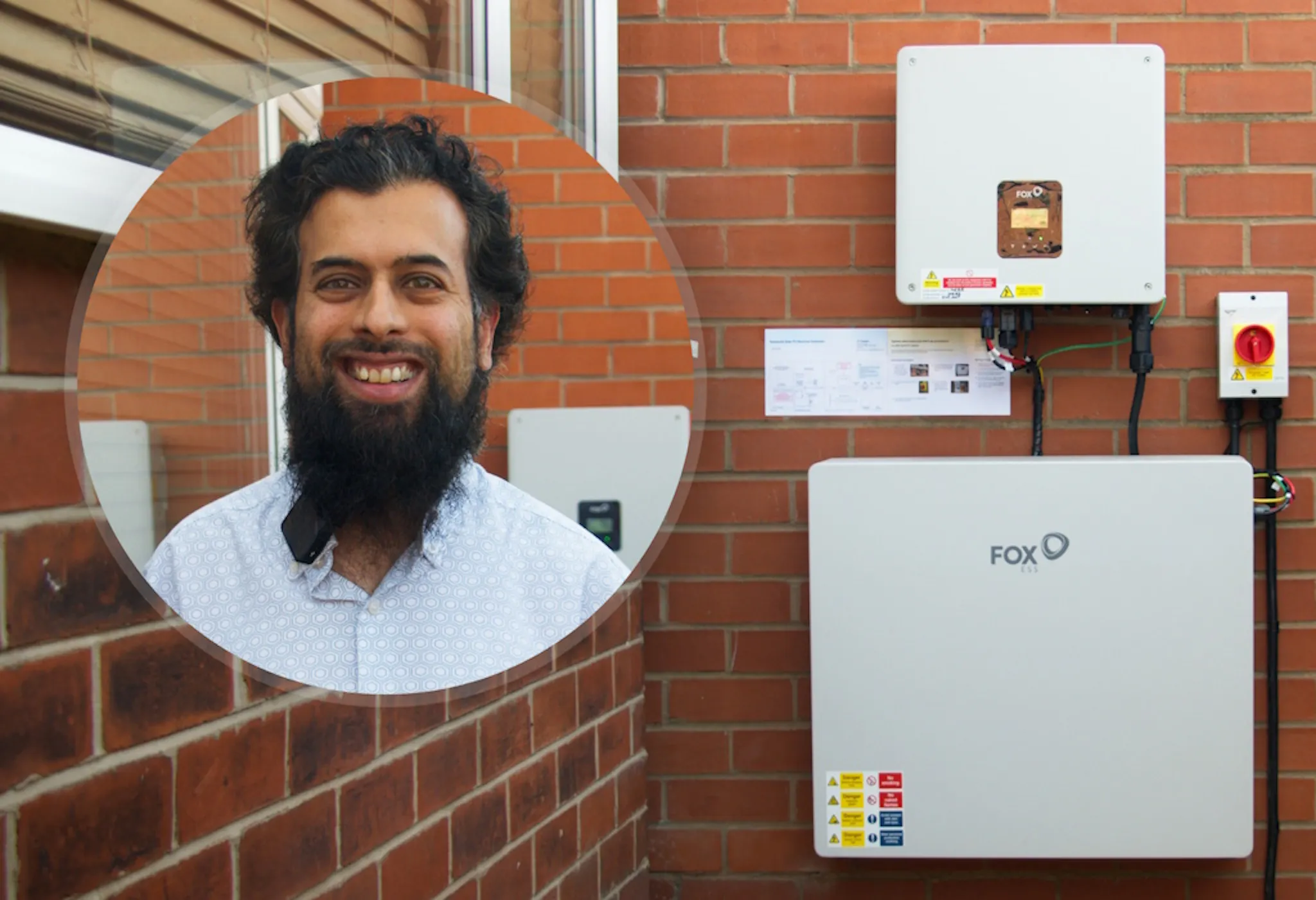 Riaz in a circle in front of his battery and inverter on a brick wall