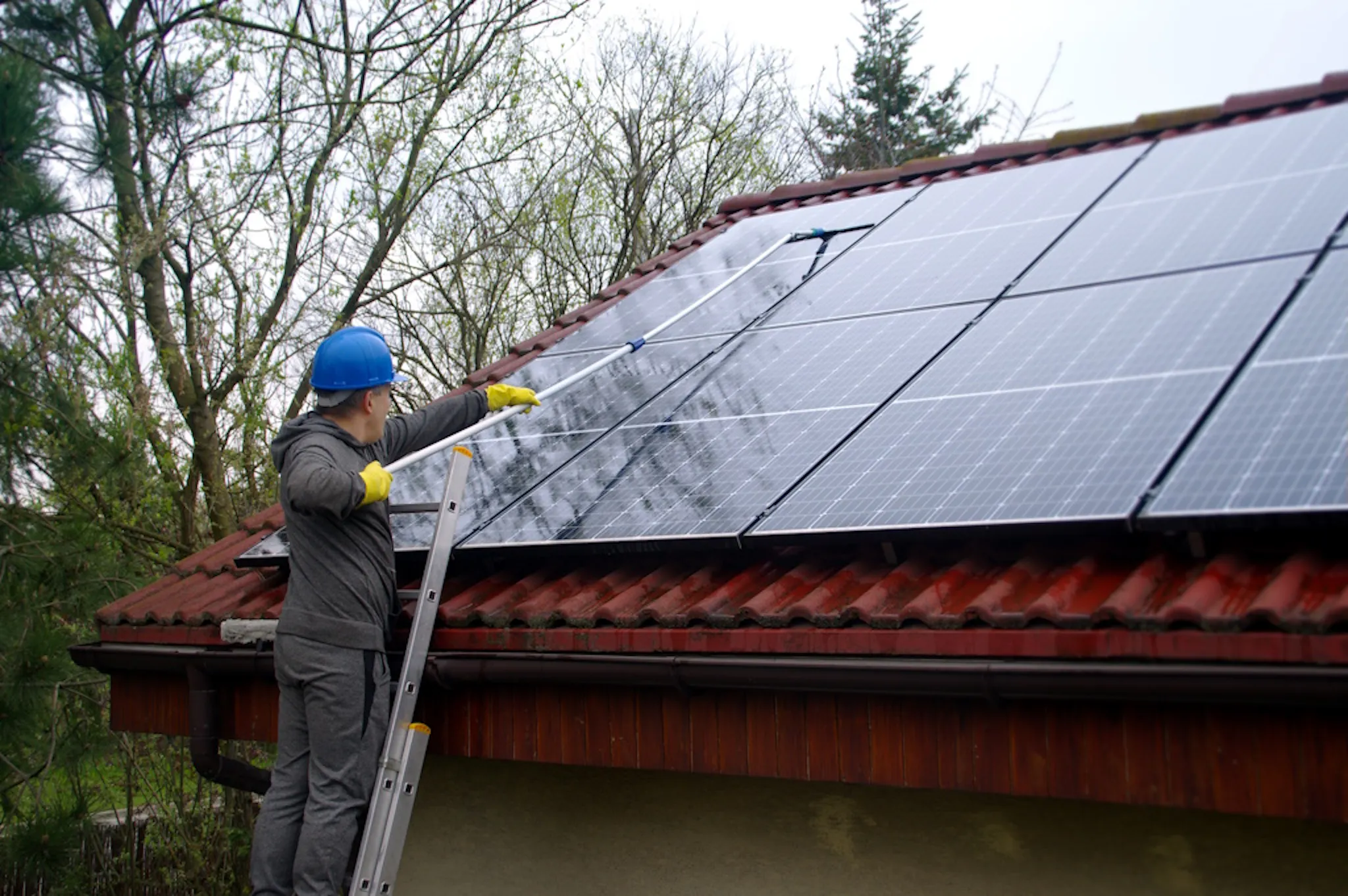 person with a brush on a pole, cleaning black solar panels on a terracotta roof