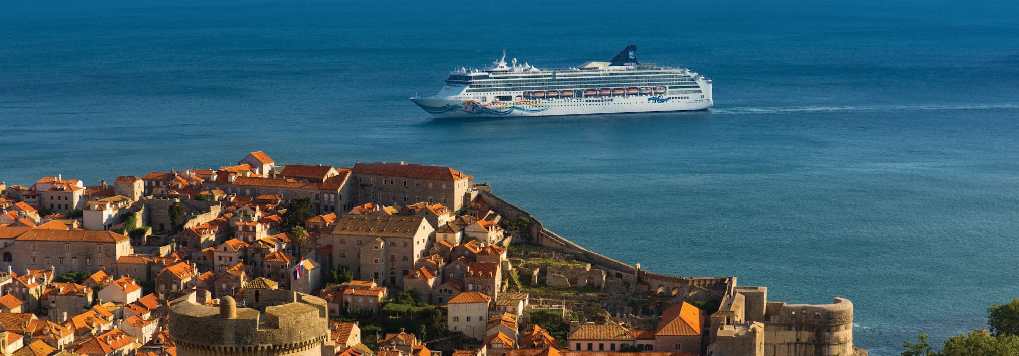 Norwegian Spirit i bakgrunden utanför en kust i Medelhavet med byggnader i sten i förgrunden.