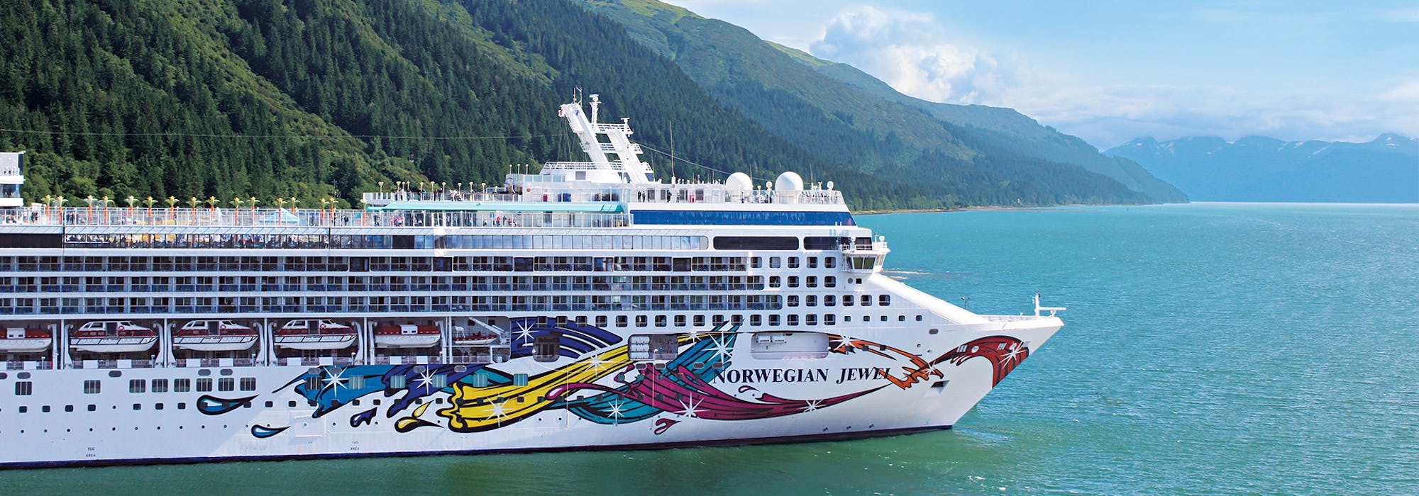 Bild på Norwegian Jewel tagen från sidan med höga berg och blått vatten runtomkring.