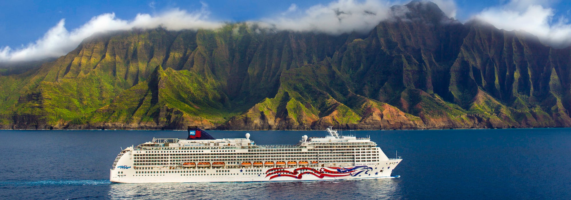 Fartyget Pride of America kryssas i det kristallblå vattnet utanför Hawaiis kust med storslagen natur och berg i bakgrunden.
