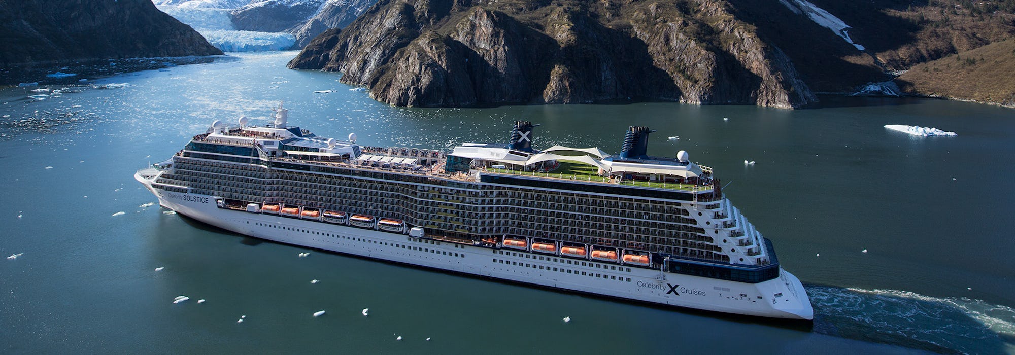 Foto på Celebrity Solstice som uppenbarar sig förgrunden med snötäckta berg i bakgrunden.