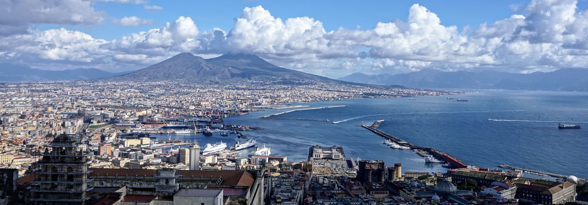 Bild från ovan på Neapels hamn med mängder av byggnader och vulkanen Vesuvius i bakgrunden.