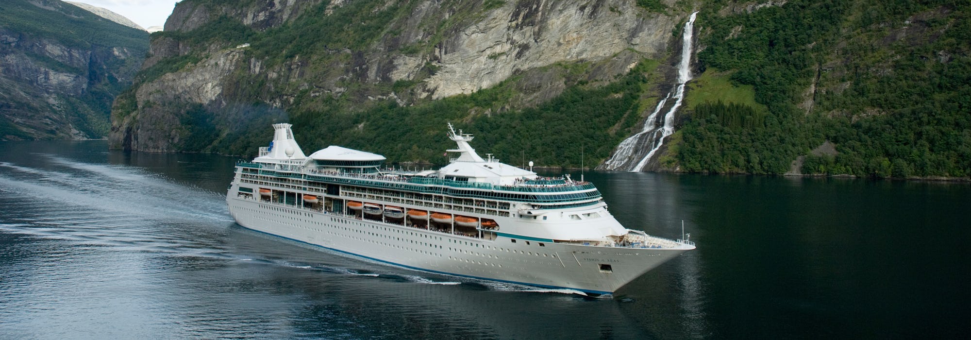 Vision of the Seas kryssar genom de storslagna norska fjordarna. I bakgrunden syns gröna berg och ett vattenfall.