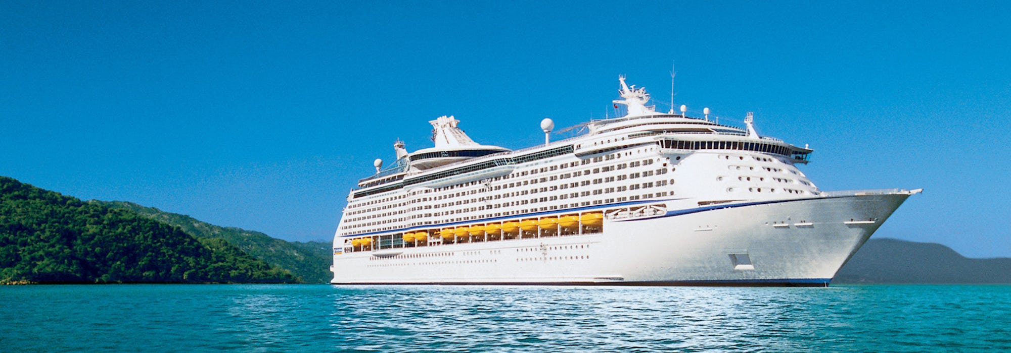 Fartyget Voyager of the Seas på håll med vackert blått vatten i förgrunden och smaragdgrön skog i bakgrunden.
