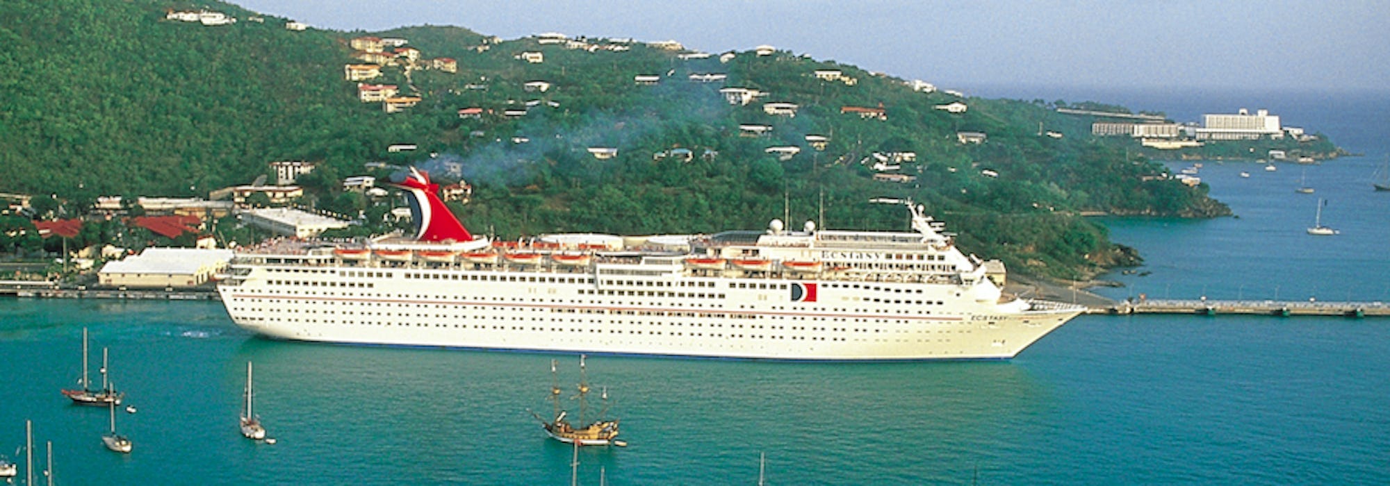 Fartyget Carnival Ecstasy ligger vid en hamn med en grön kulle i bakgrunden och mindre segelbåtar i framkant.