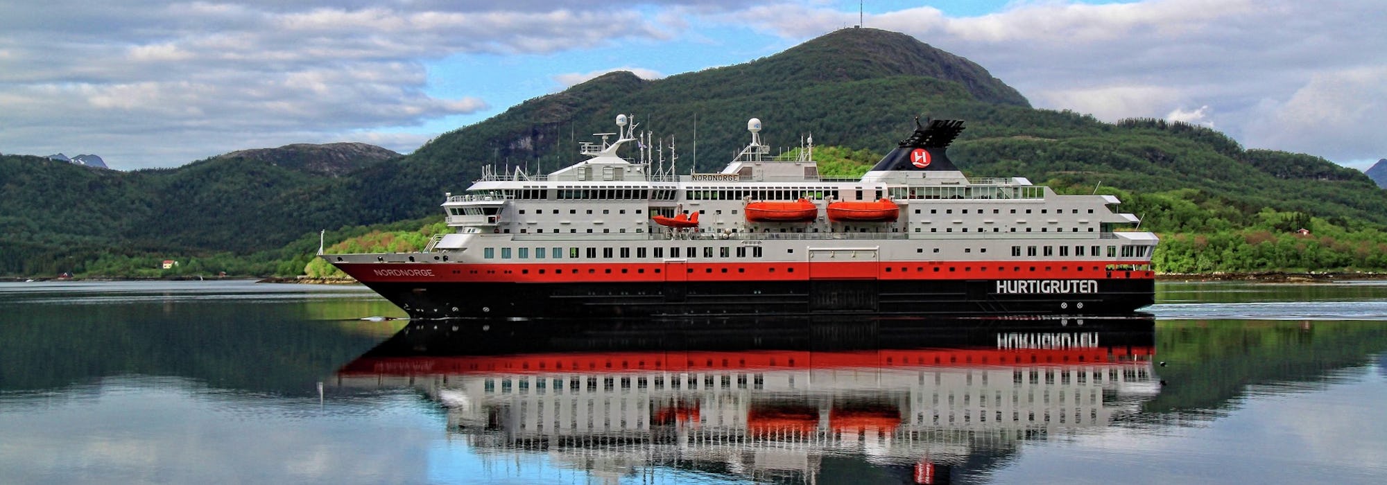 Bild från sidan på Hurtigrutens fartyg Ms Nordnorge som stilla glider fram i vattnet med gröna berg i bakgrunden.