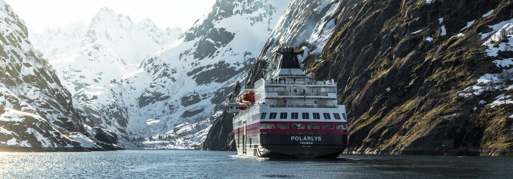 Bild på fartyget MS Polarlys akter medan fartyget lider fram mellan snötäckta berg i de norsk fjordarna.