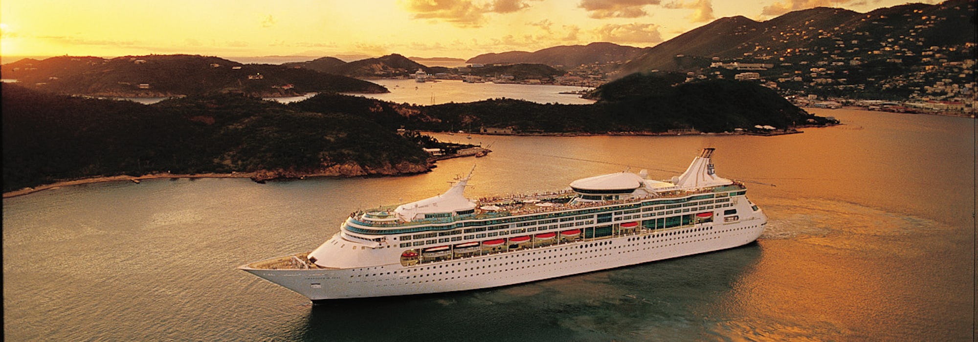Bild på Rhapsody of the Seas, taget från sidan, utanför en Karibisk ö i solnedgången.