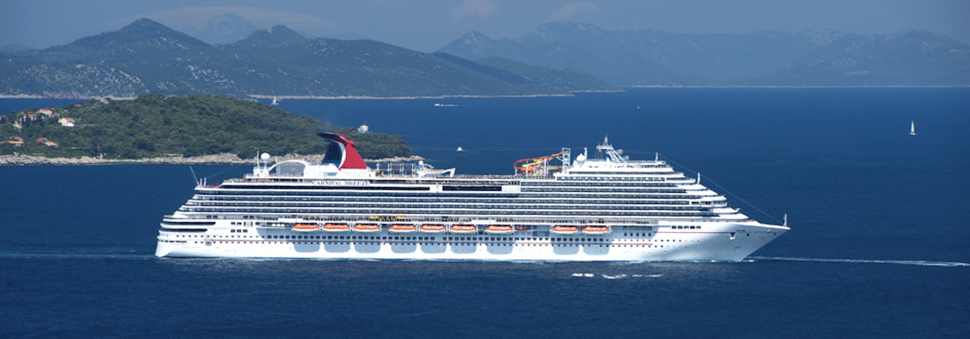Bild på Carnival Breeze med en grön ögrupp och höga berg i bakgrunden.