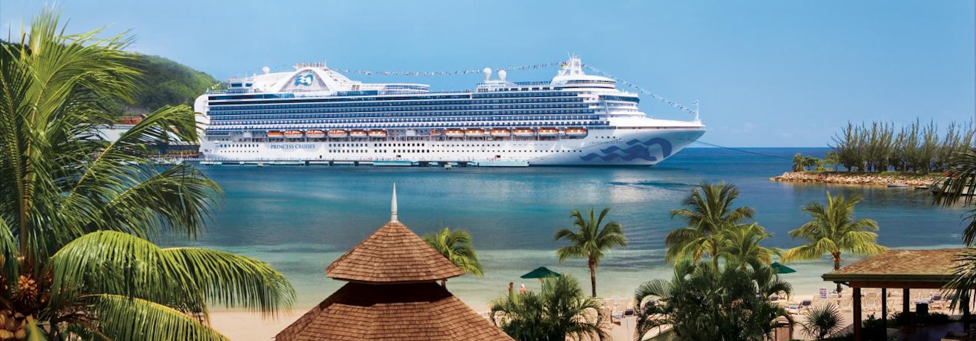 Fartyget Crown Princess ligger ankrad utanför en hamn i exotiska Jamaica. I framkant syns palmer och vit sand.