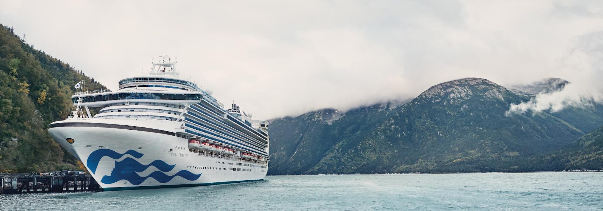 Fartyget Ruby Princess i framkant och höga berg med moln som omringar dem i bakgrunden.