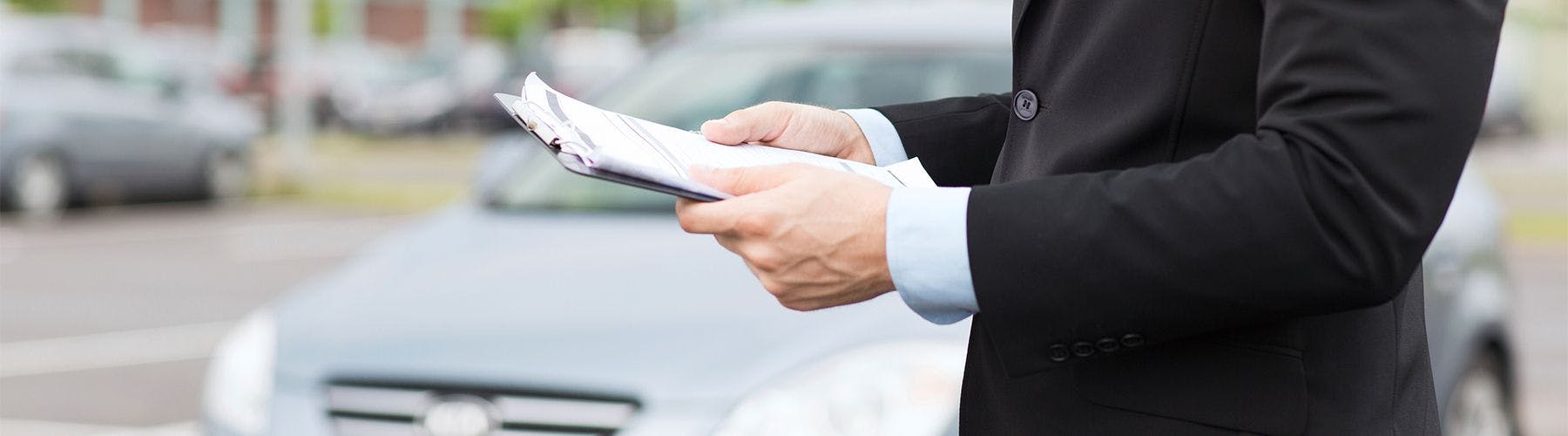 Person holding a clipboard in front of a blurred grey car 