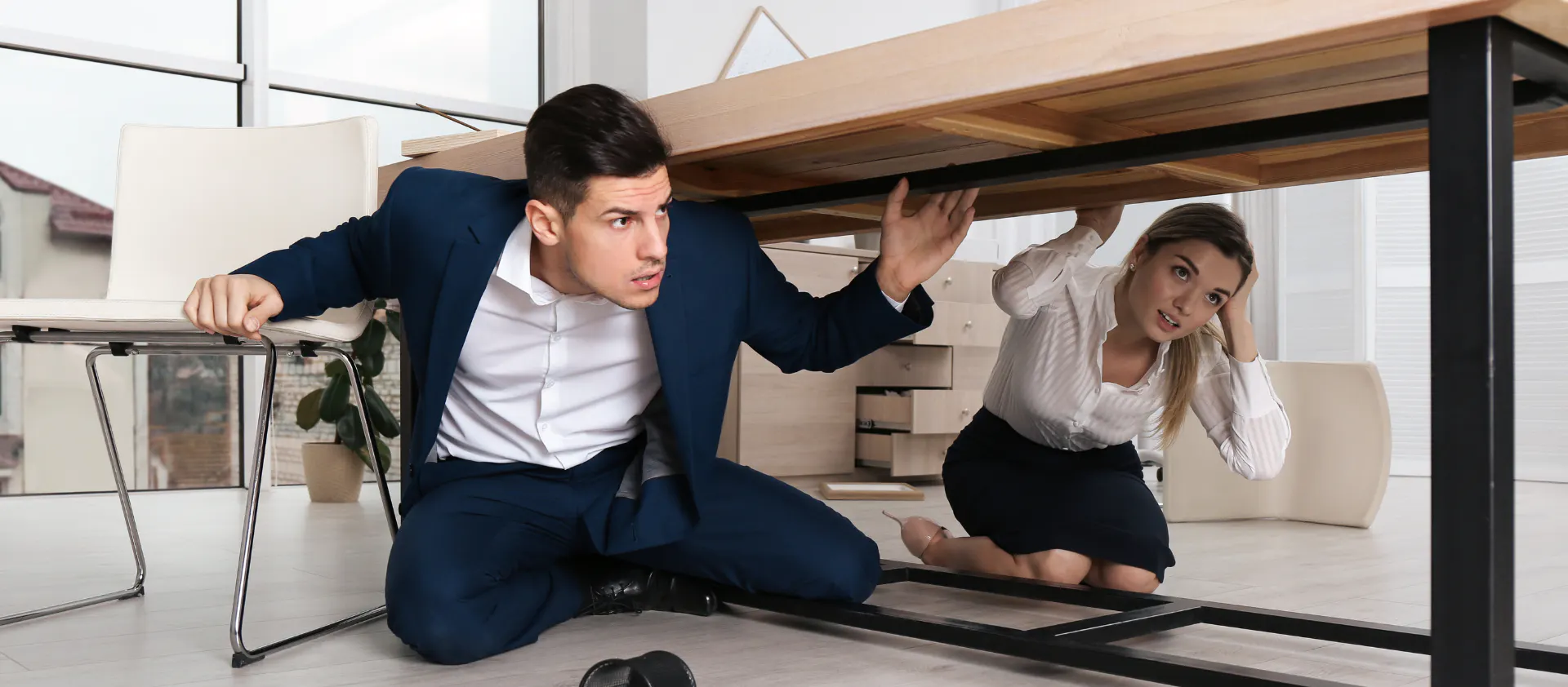 Dos personas se protegen debajo de una mesa durante un sismo o terremoto, manteniéndose agachadas y cubriendo su cabeza mientras esperan que pase el movimiento.