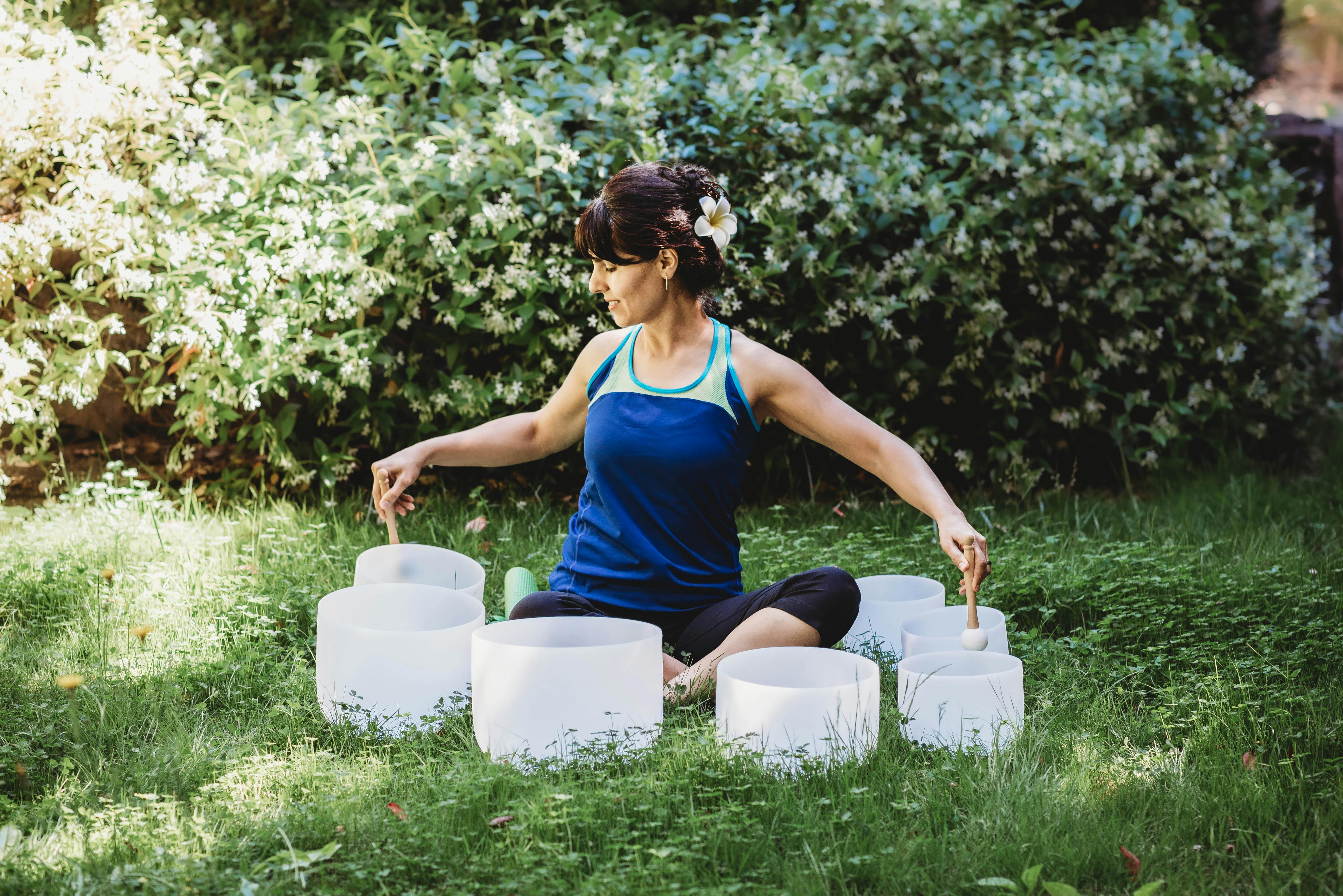 Woman playing crystal bowls while sitting in the grass in front of a set of jasmin bushes that are in bloom.