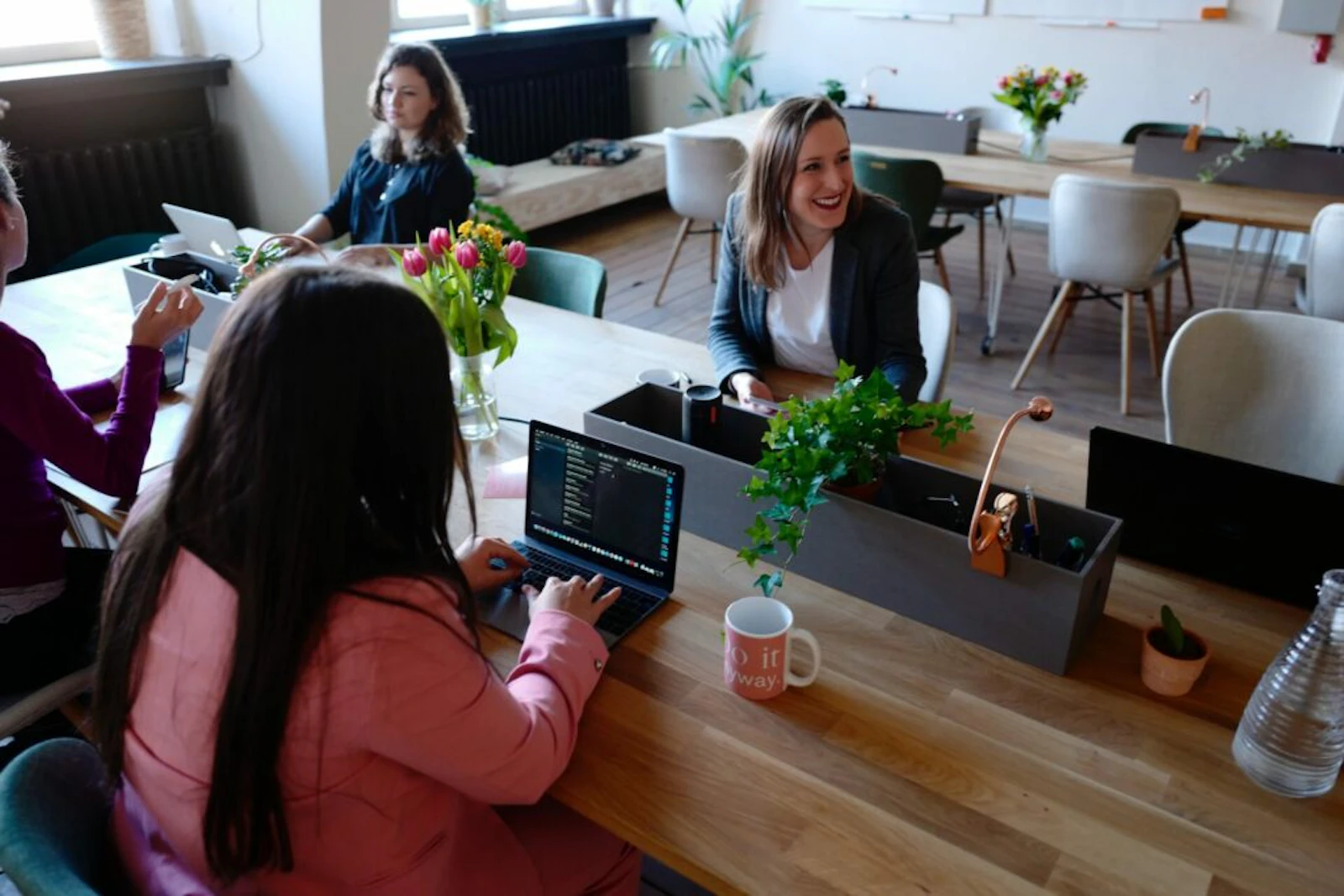 Women working independently on laptops at a shared coworking table with plants