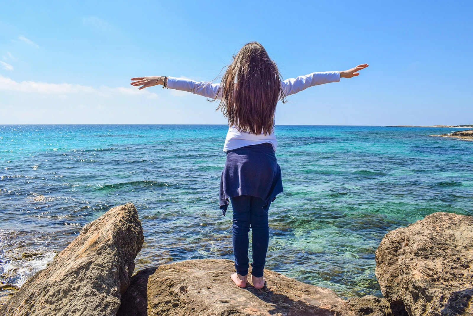 A woman standing on top of a rock overlooking the sea with her hands spread