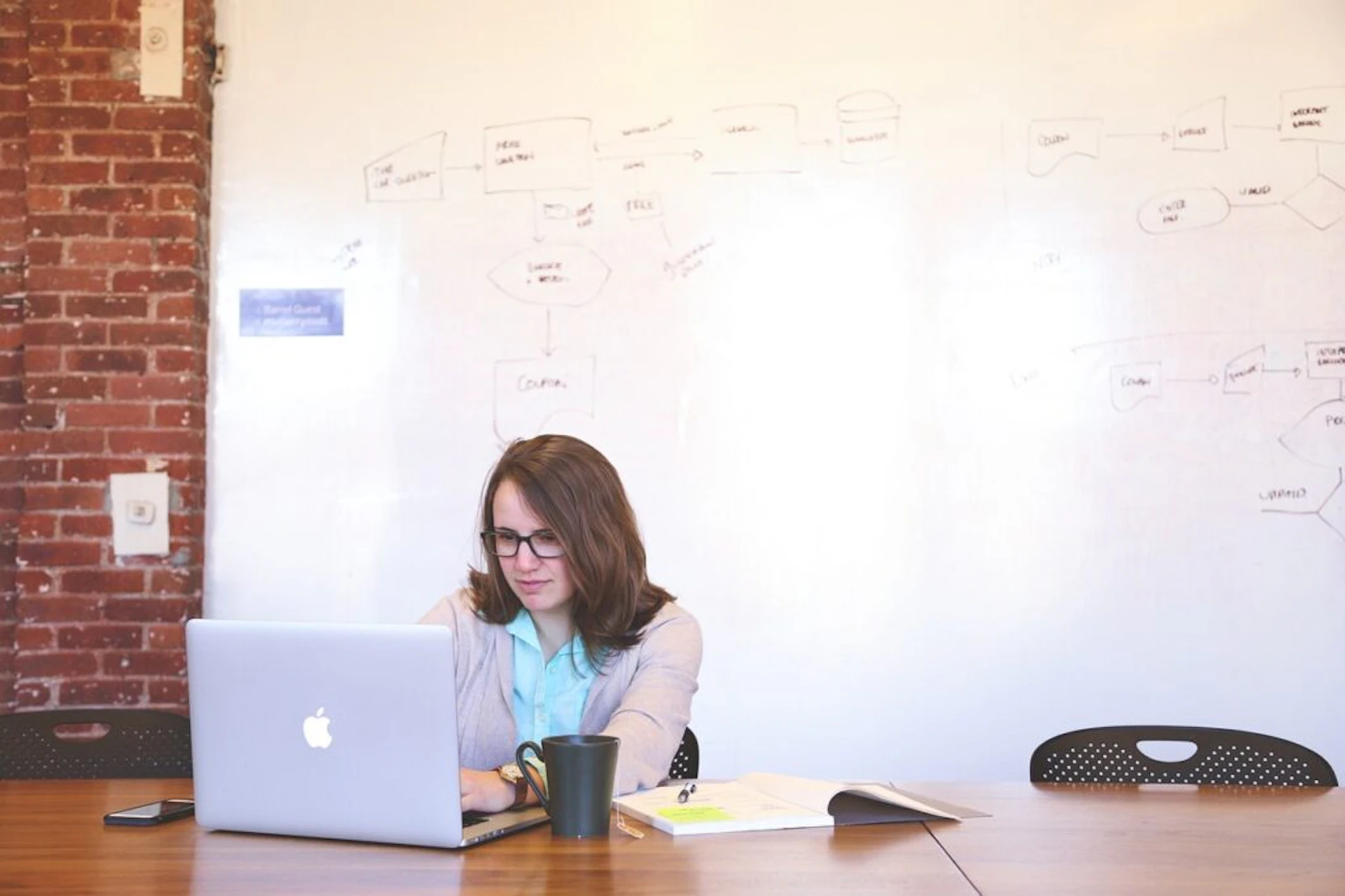 Woman working on a laptop at a desk in a loft-style office with exposed brick wall behind her