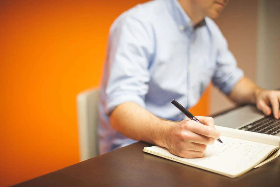 a man working on a laptop and taking notes in a notebook