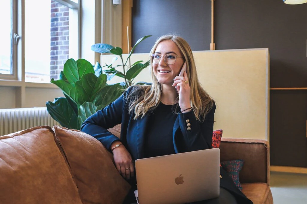 Smiling woman talking on the phone with a laptop in a bright, relaxed office setting