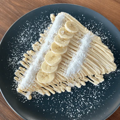Palačinka sa belim mlečnim kremom, bananom i kokosom na tamnom tanjiru