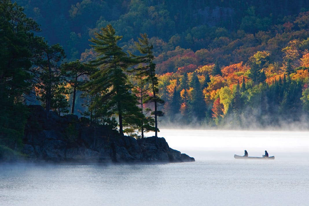 Two people on a canoe, cutting through a foggy lake towards a rocky shore dotted with evergreens. Fall-coloured trees in the background.