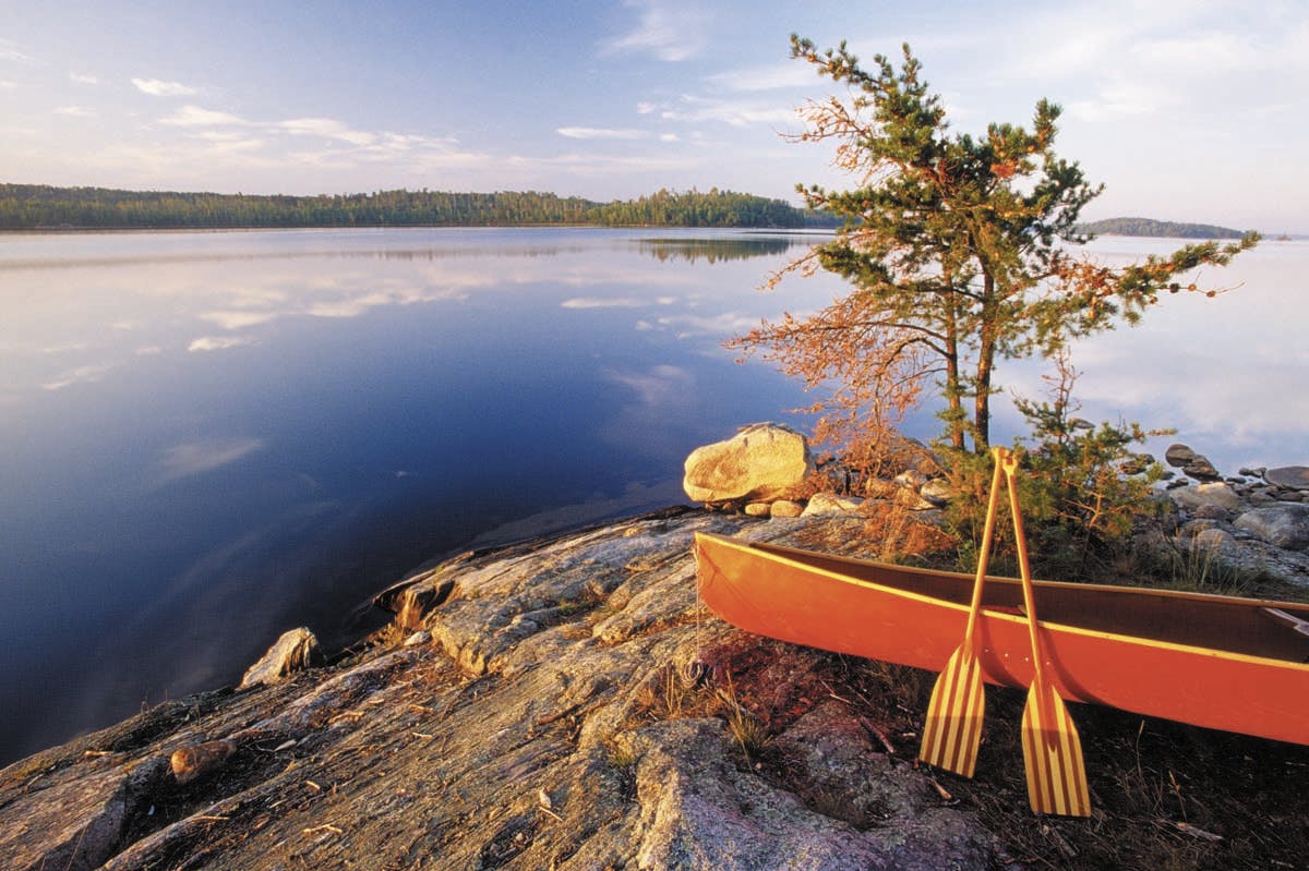 Bright red canoe and wooden paddle laid on a rocky shoreline overlooking a clear blue lake. Green trees dot the shoreline in the distance.