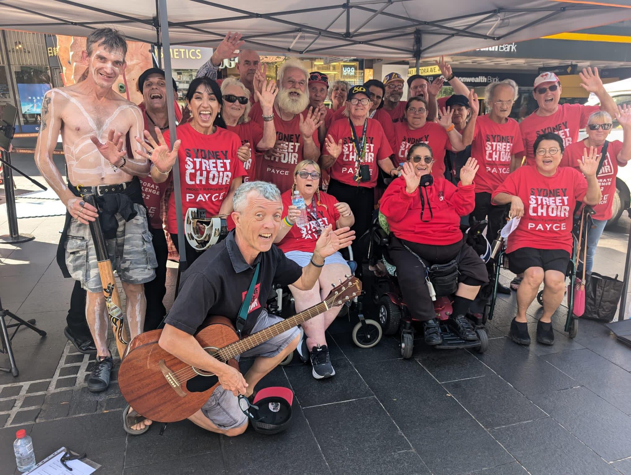 The Sydney Street Choir standing/kneeling/sitting under an awning for the Stronger Together event at Chatswood Mall