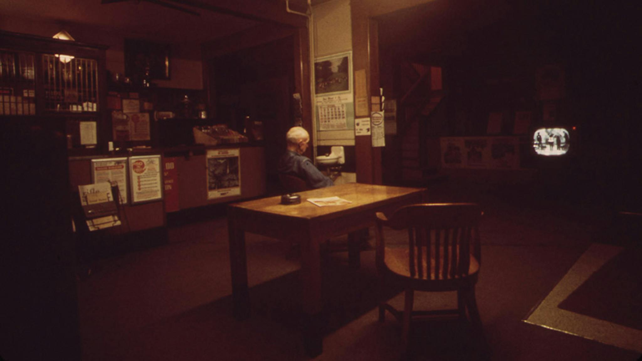 Photo of an older man with white hair sitting alone at a table. He looks away from the camera.