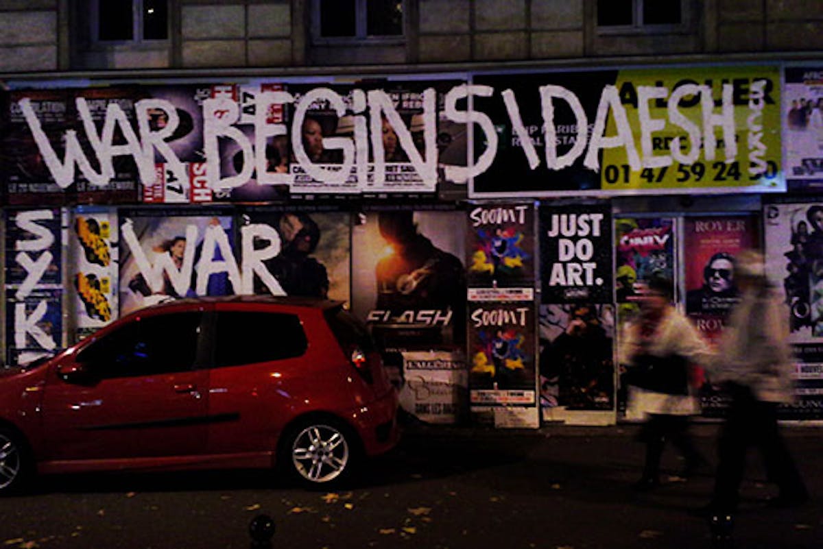 'WAR BEGINS\DAESH SUCKS' is graffitied in block letters in white paint over the top of advertising posters in a city street 