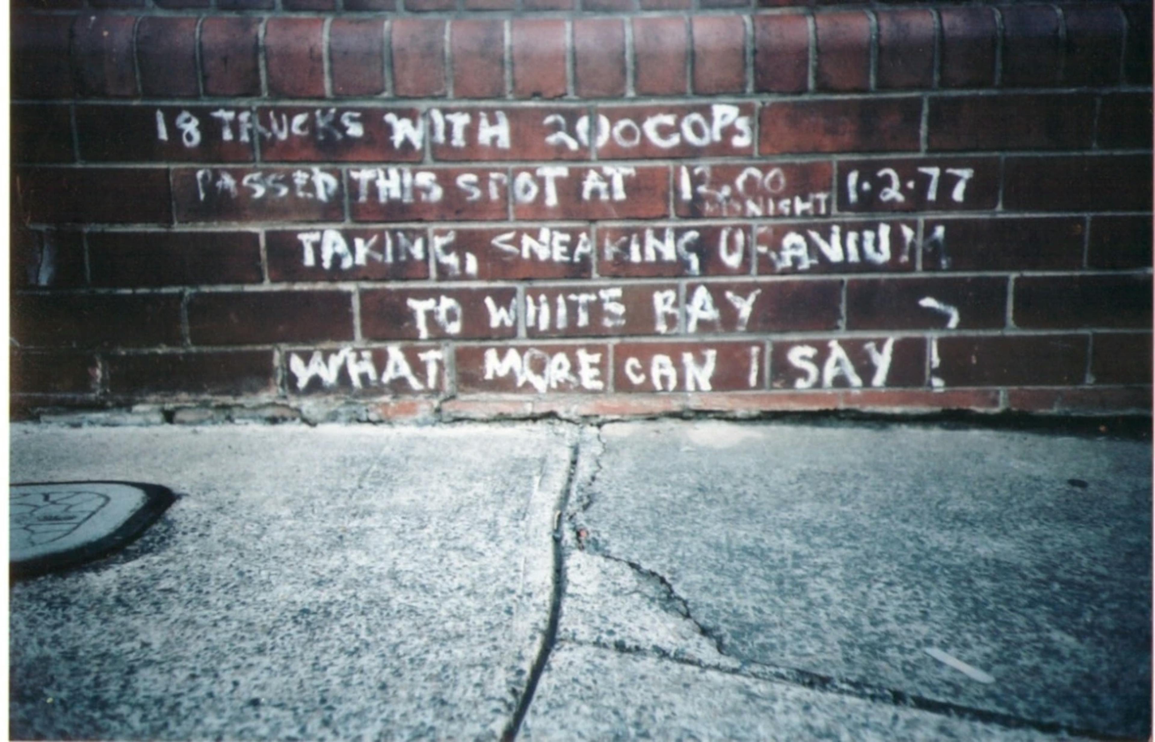 A brick while graffitied with white text: 18 TRUCKS WITH 200 COPS PASSED THIS WAY AT 12.00 MIDNIGHT 1.2.77 TAKING, SNEAKING, URANIUM  TO WHITE BAY WHAT MORE CAN I SAY?'