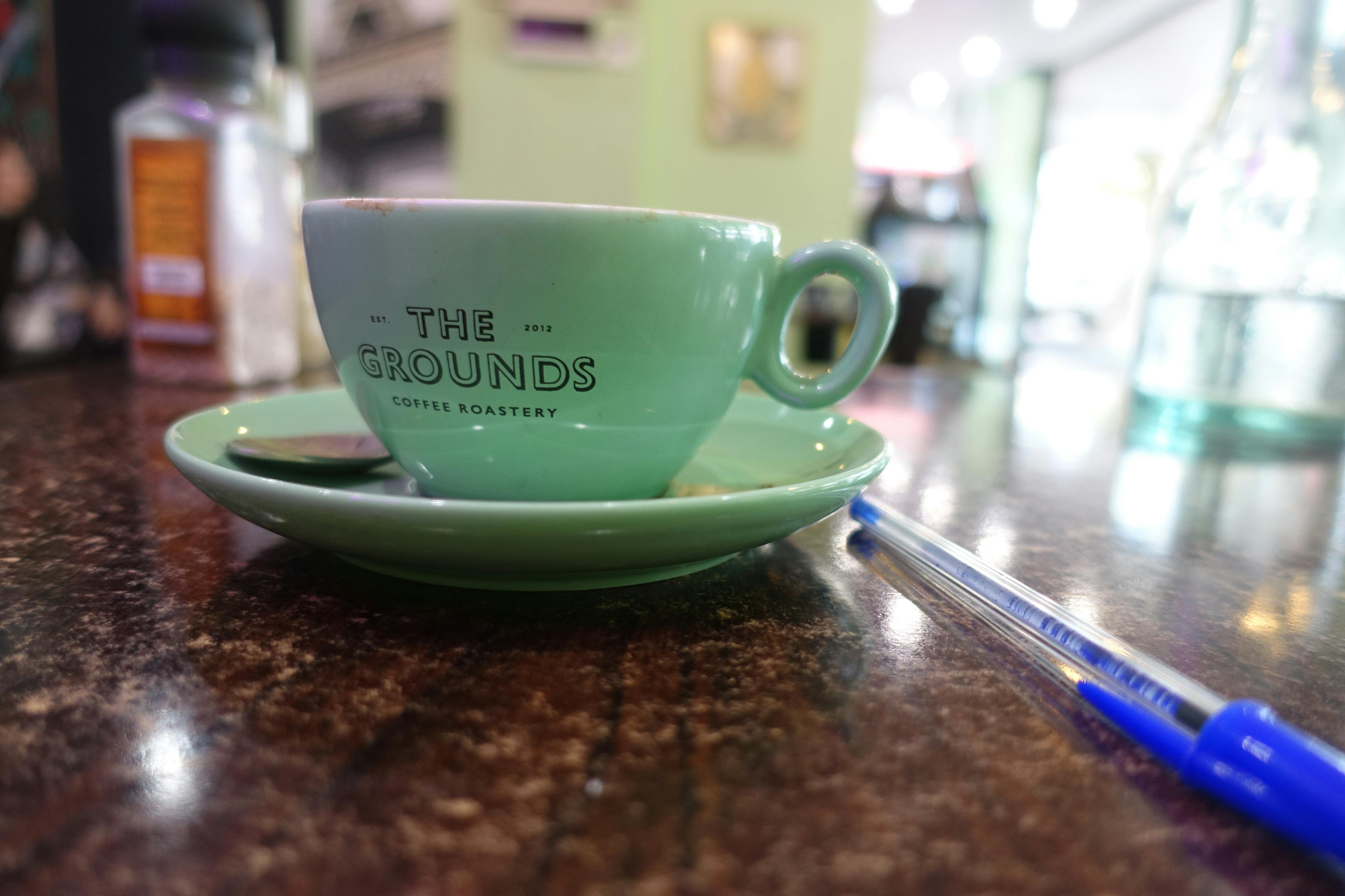 A mug and saucer with ‘The Grounds Coffee Roastery’ printed on the mug, and a pen on a wooden table. 